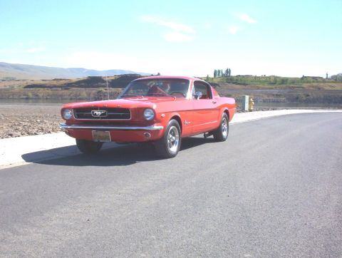 1965 Ford Mustang - A red 1965 Ford Mustang coupe is parked on a paved road with a body of water and hills in the background.
