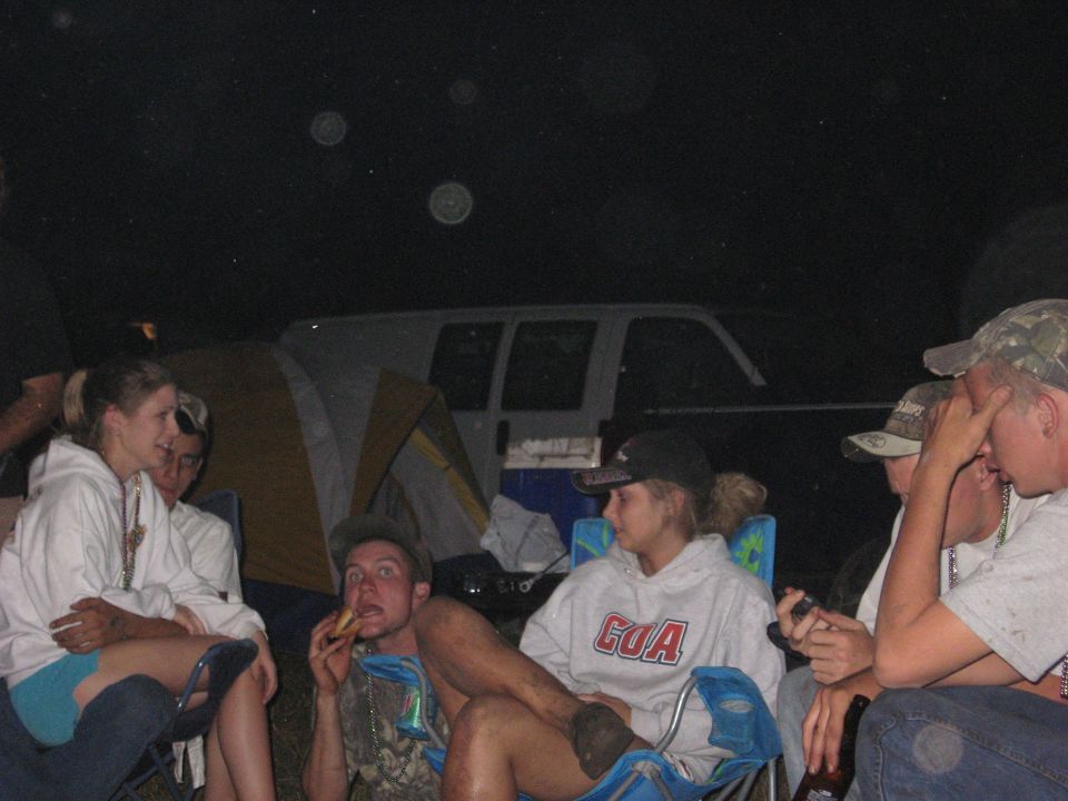 Group of young adults sitting around a campfire at night, with a tent and a van visible