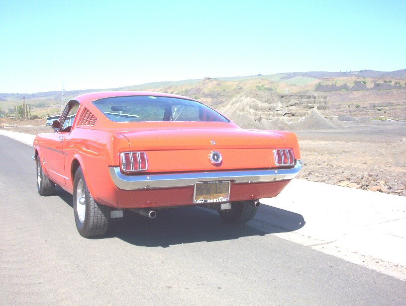 Orange 1965 Ford Mustang fastback, rear three-quarter view, parked beside a road in arid hilly terrain