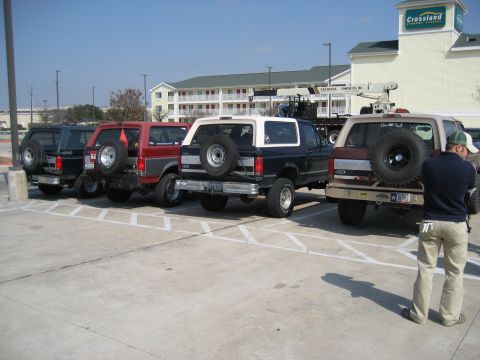 1986 Ford Bronco - Multiple Ford Broncos are parked in a lot, with a building in the background and a person standing near one of the vehicles.
