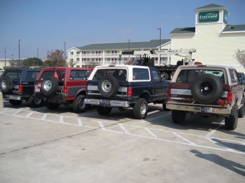 1986 Ford Bronco - A lineup of Ford Broncos, including a tan 1987 model, are parked in a lot outside a hotel.