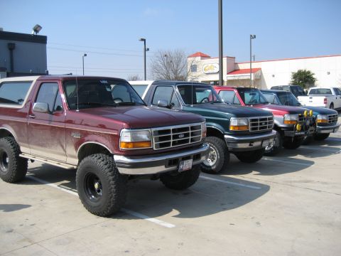 1986 Ford Bronco - Several trucks, including a maroon Ford Bronco, are parked in a lot on a sunny day.