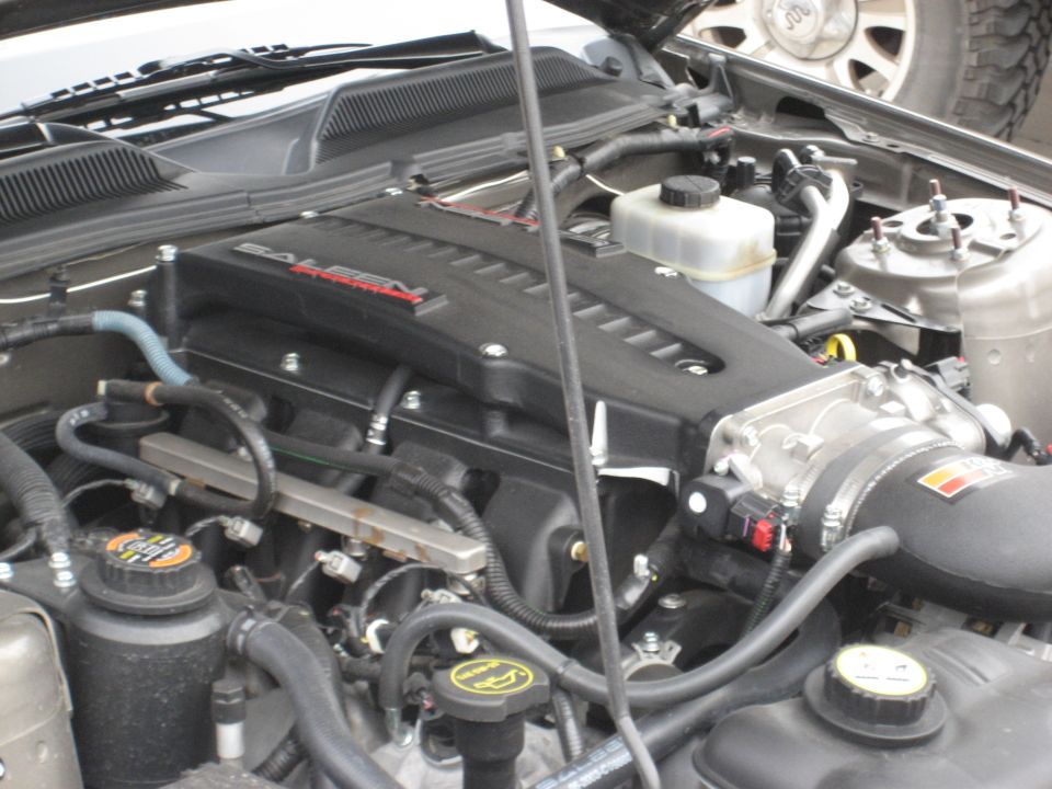 Engine bay with black intake manifold labeled Saleen, hoses, and fluids. A tire is visible in the upper right