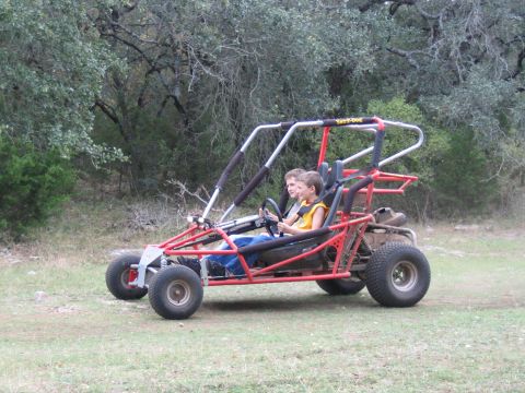 1996 Ford Bronco - Two boys ride in a red go-kart outdoors, with a wooded area behind them.