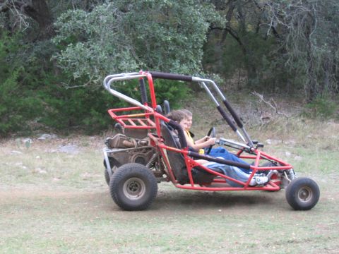 1996 Ford Bronco - A red dune buggy carries two children through a wooded, grassy area.