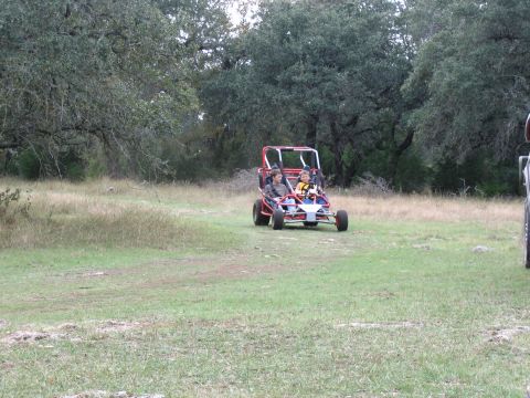 1996 Ford Bronco - A dune buggy with two occupants drives across a field with trees in the background.