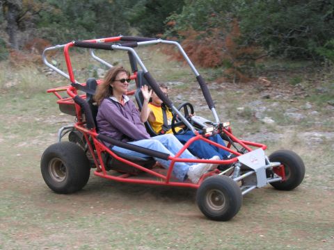 1996 Ford Bronco - A woman and child wave from a red dune buggy as they drive on a dirt path through a wooded area.