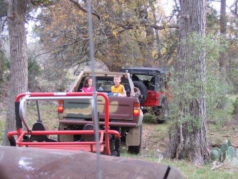 1996 Ford Bronco - Two off-road vehicles, a red Jeep and a camouflaged pickup truck, are parked among trees with children riding in the back of the truck.