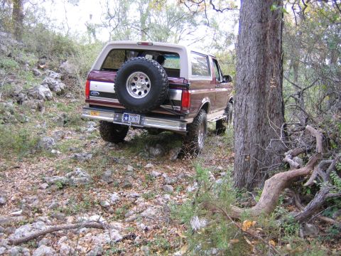 1996 Ford Bronco - A maroon 1996 Ford Bronco navigates a rocky, wooded terrain, its spare tire prominently displayed on the rear.