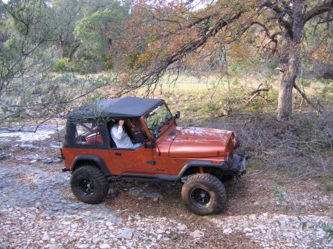 1996 Ford Bronco - An orange Jeep Wrangler is parked off-road on a rocky surface with trees and brush surrounding it.