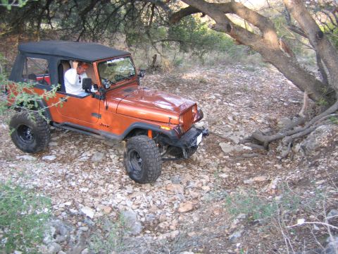 1996 Ford Bronco - Orange Jeep Wrangler navigates a rough, rocky terrain under a tree canopy.