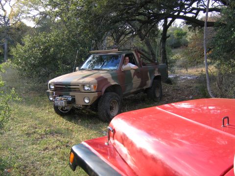 1996 Ford Bronco - A camouflage pickup truck is parked off-road, with a red vehicle partially visible in the foreground.
