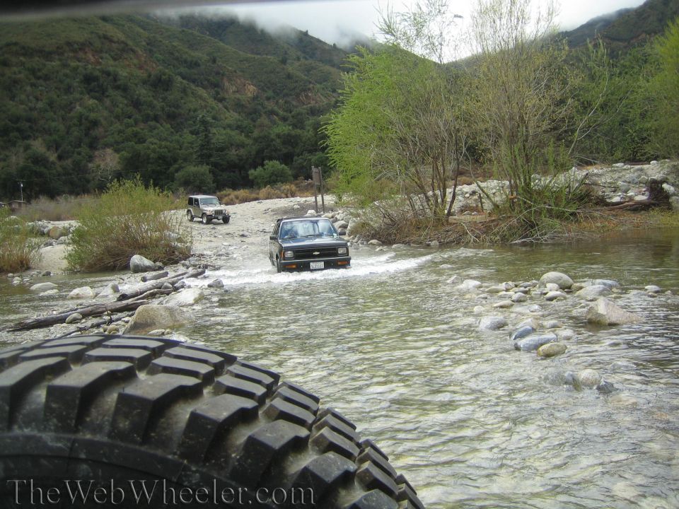 Two SUVs driving through a rocky riverbed with forested mountains in the background. A large tire tread is visible in the foreground