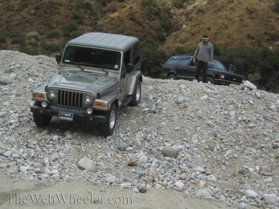 Gold Jeep Wrangler parked on a rocky hill with a man standing next to a black Chevrolet Blazer behind it