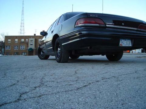 1992 Ford Crown Victoria - The rear of a black 1992 Ford Crown Victoria is shown from a low angle, with a building and tower in the background.