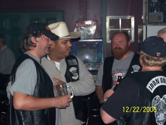 Men in leather vests and hats talking in a dimly lit bar with a pinball machine in the background