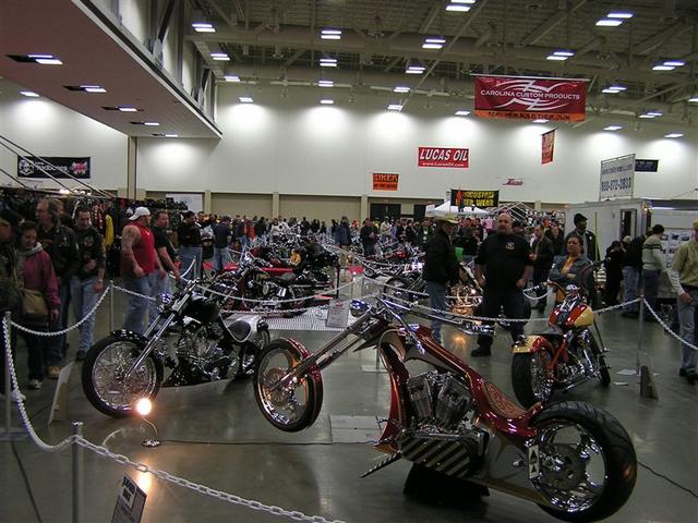 Custom choppers and motorcycles at a show, with people milling around. One red and chrome chopper is in the foreground