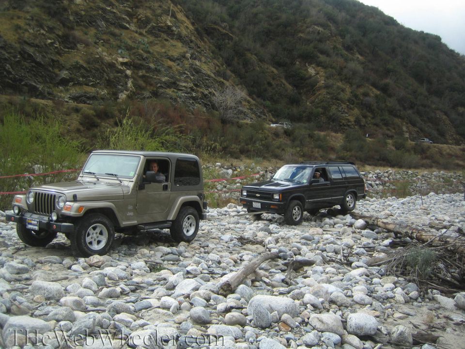Two SUVs, a tan Jeep Wrangler and a black Chevrolet S-10 Blazer, drive on a rocky riverbed with a mountain in the background