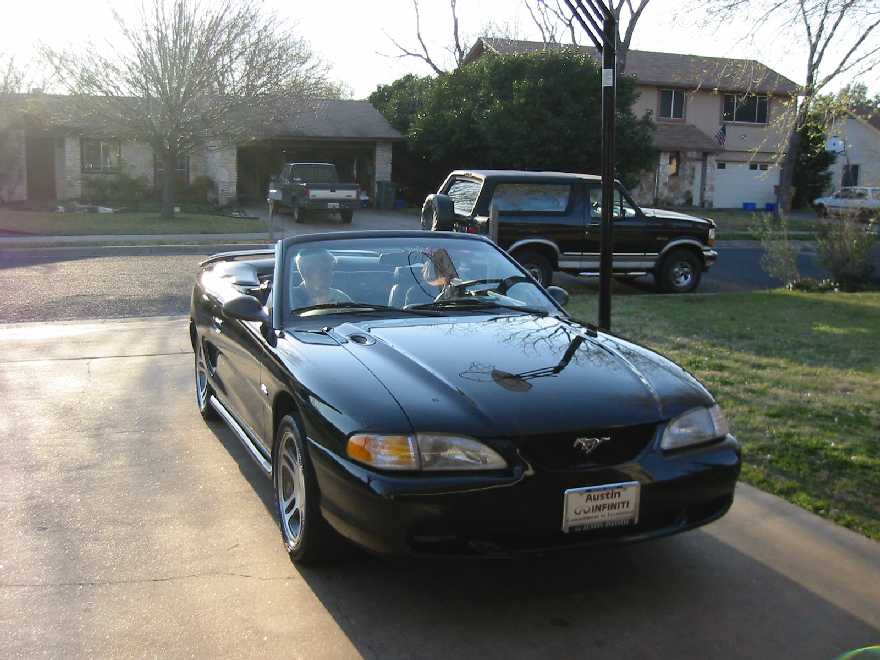 Black convertible Ford Mustang parked on driveway, front 3/4 view. A Ford Bronco is parked in the background
