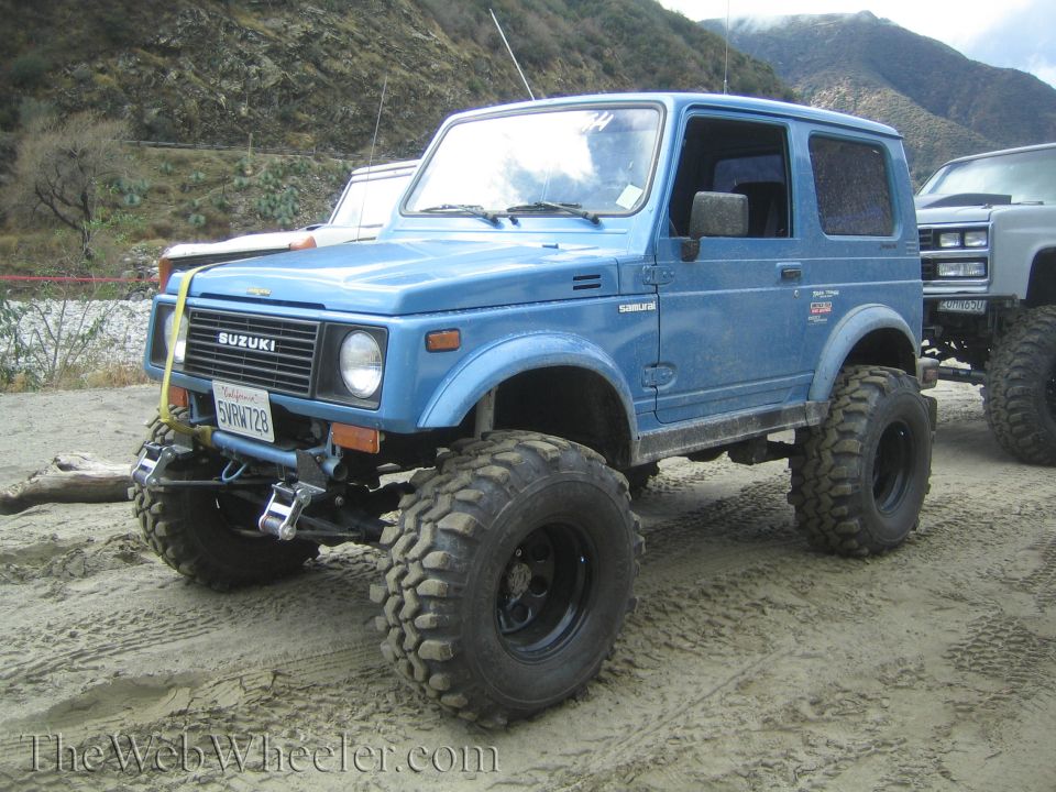 Blue 1987 Suzuki Samurai with large off-road tires parked on sand, another vehicle to the right