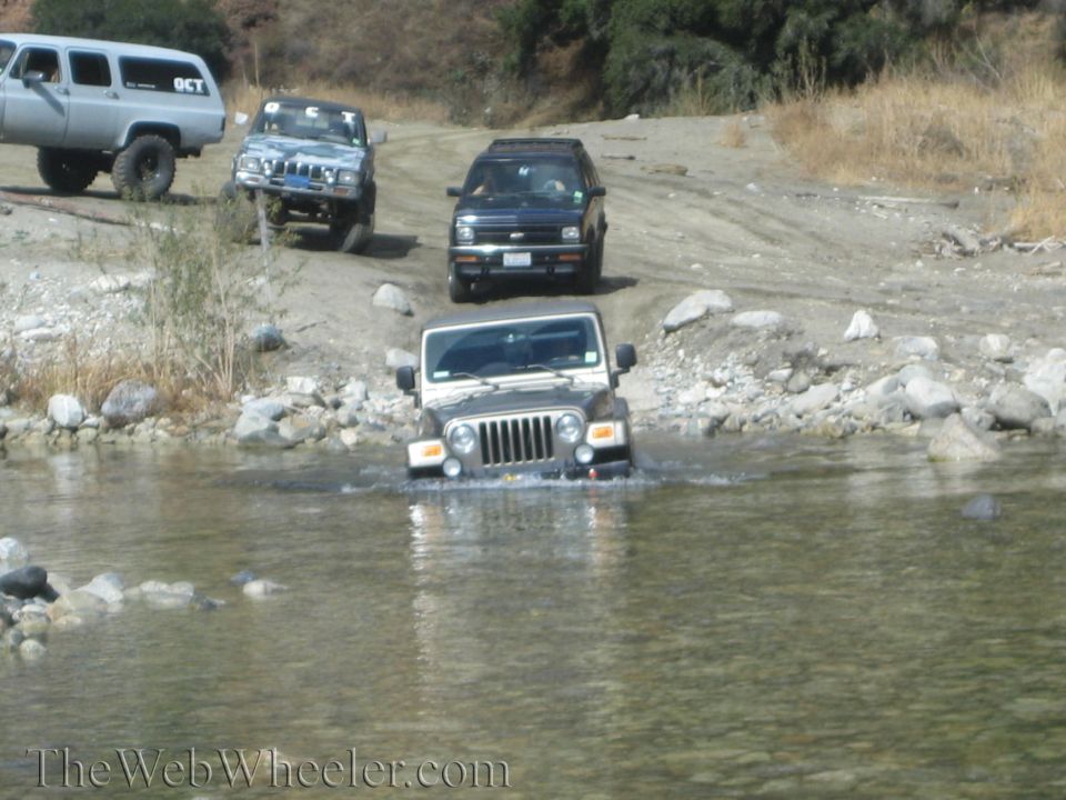 Gray Jeep Wrangler driving through shallow water, front view. Other SUVs are visible on the dirt path behind it