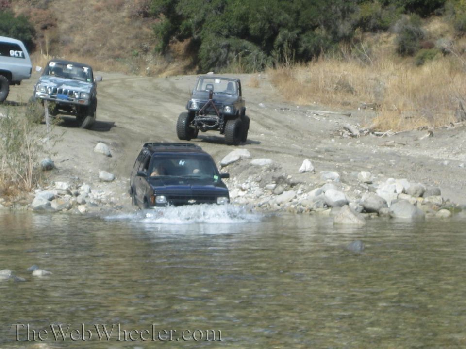 Black SUV driving through water, with a Jeep and a Suzuki Samurai behind it on a dirt road