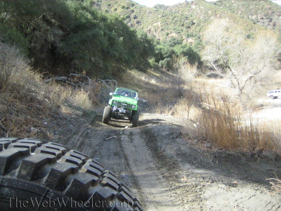 Green off-road vehicle climbing a dirt trail. Another white vehicle is parked in the background