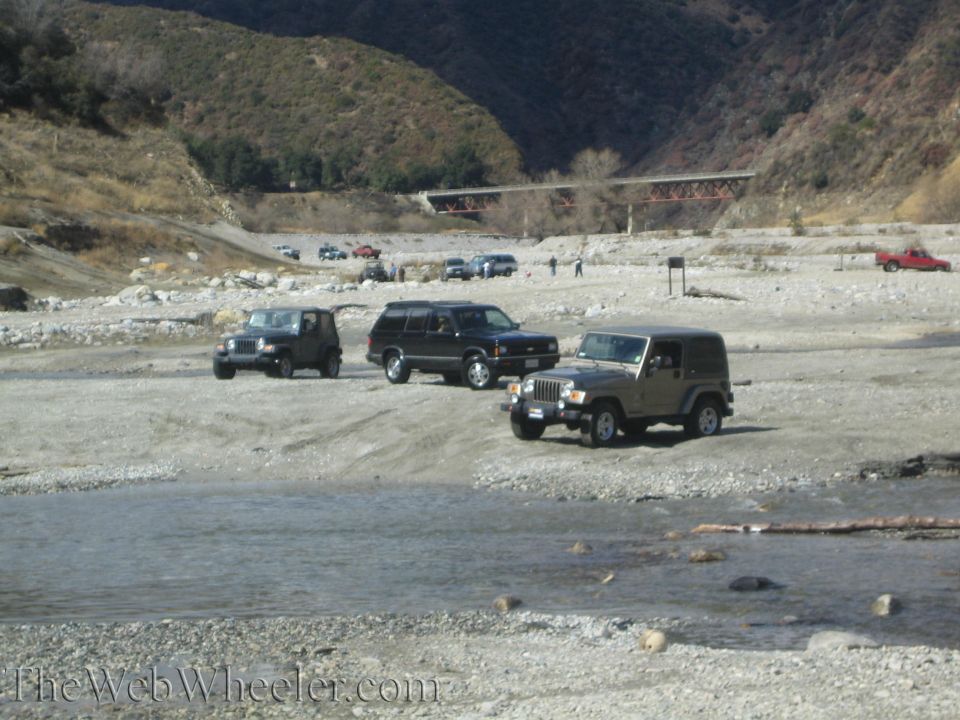 Tan Jeep Wrangler parked on a rocky riverbed with a black Jeep and black SUV nearby