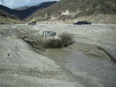 1987 Suzuki Samurai - A Jeep Wrangler kicks up a large spray of mud while navigating a water-filled path in a dry, mountainous area.