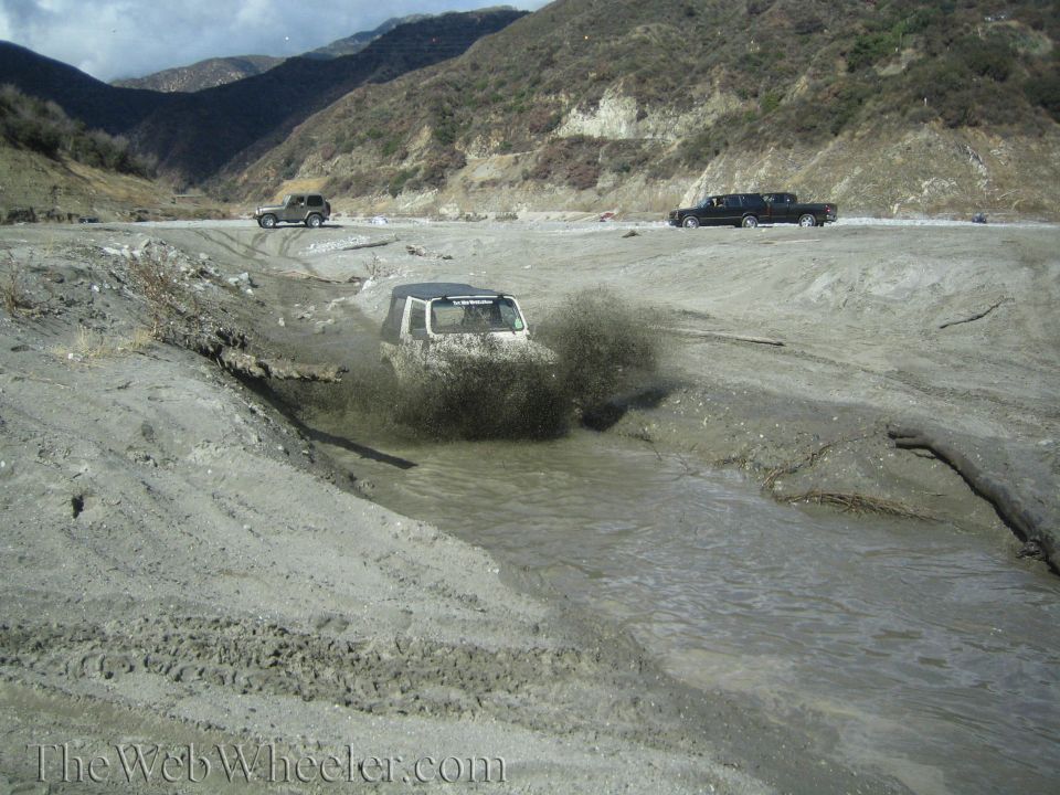 Jeep Wrangler driving through a muddy stream in a canyon. Another SUV and a black sedan are visible further back