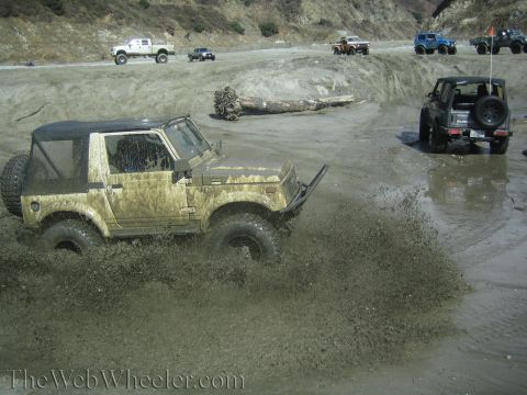 1987 Suzuki Samurai - Multiple off-road vehicles navigate a muddy terrain, with one Suzuki Samurai kicking up a large spray.