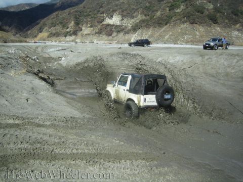 1987 Suzuki Samurai - A white 1987 Suzuki Samurai navigates a muddy terrain, kicking up spray as it goes.