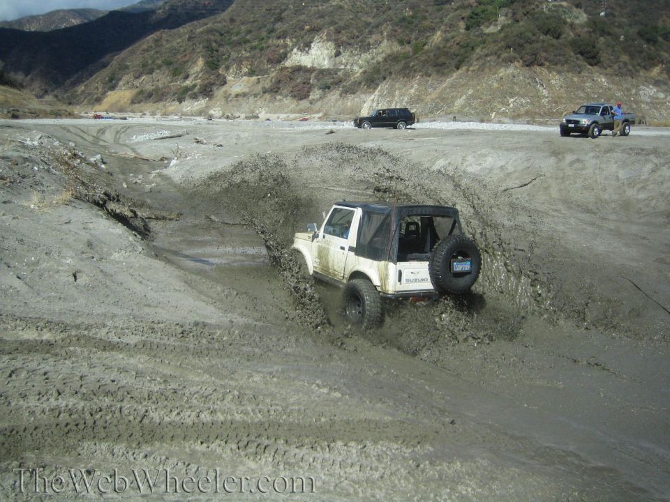 White 1987 Suzuki Samurai with soft top, driving through mud, rear view, spare tire visible