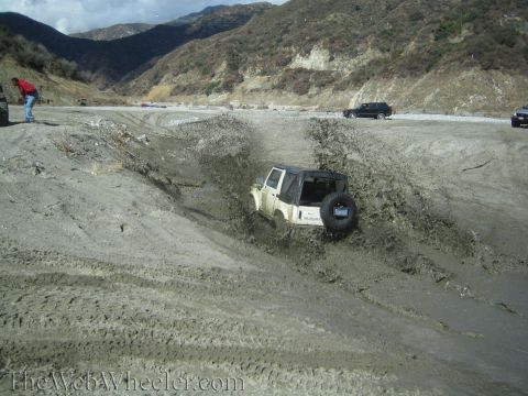 1987 Suzuki Samurai - A white 1987 Suzuki Samurai navigates a muddy terrain, kicking up a spray of dirt and water. Other vehicles are visible in the distance.