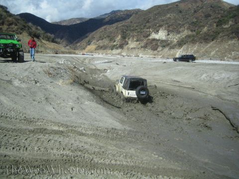 1987 Suzuki Samurai - Multiple off-road vehicles navigate a muddy, dry riverbed with steep, barren hillsides under a cloudy sky.