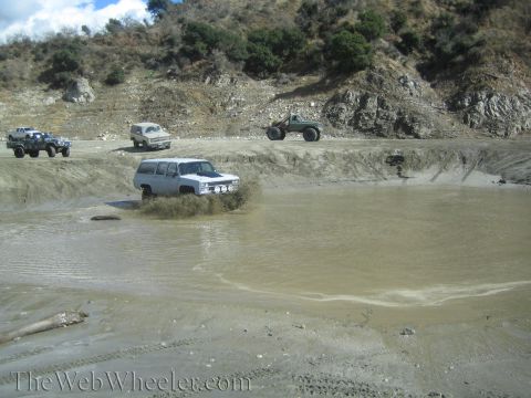 1987 Suzuki Samurai - Several off-road vehicles navigate a muddy terrain, with one white SUV prominently splashing through a water hazard.