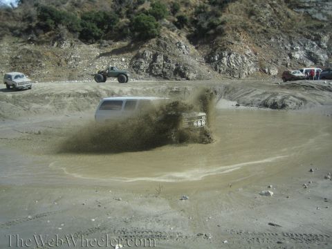 1987 Suzuki Samurai - Multiple vehicles are seen off-roading in a muddy terrain with a white SUV creating a large splash.