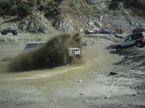 1987 Suzuki Samurai - Multiple off-road vehicles navigate a muddy track, kicking up large plumes of water and dirt.