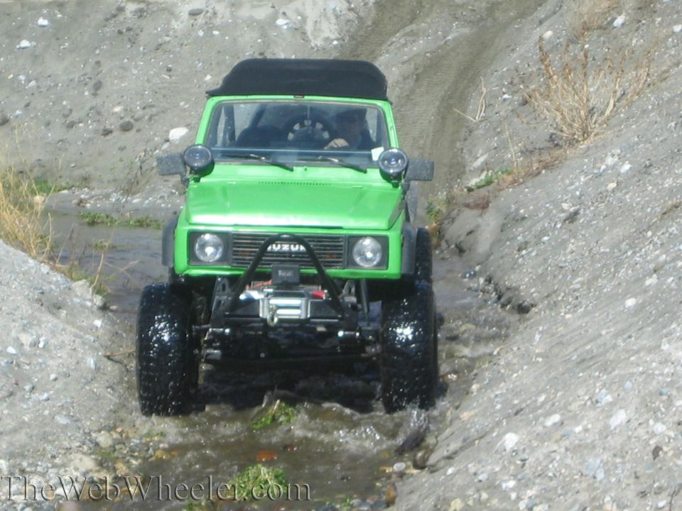 Green 1987 Suzuki Samurai driving through shallow water and dirt