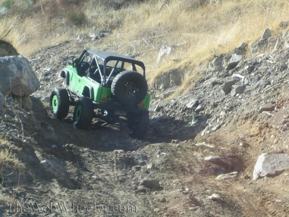 Green Jeep Wrangler with roll cage and spare tire on the back driving on a rocky trail