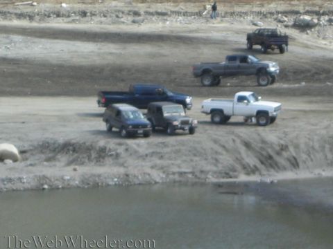 1987 Suzuki Samurai - Multiple trucks and SUVs are parked on a dirt lot next to a body of water.