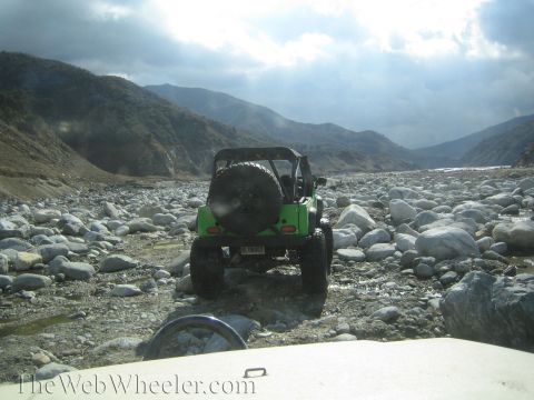 1987 Suzuki Samurai - A green off-road vehicle navigates a rocky riverbed under a cloudy sky.