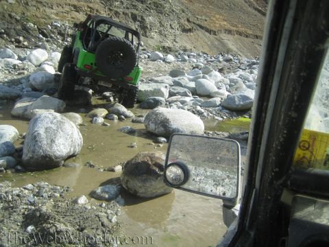 1987 Suzuki Samurai - A green Jeep navigates a rocky, shallow riverbed, with another vehicle's perspective visible.
