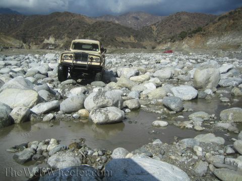 1987 Suzuki Samurai - A tan off-road vehicle navigates a rocky terrain with mountains in the background.