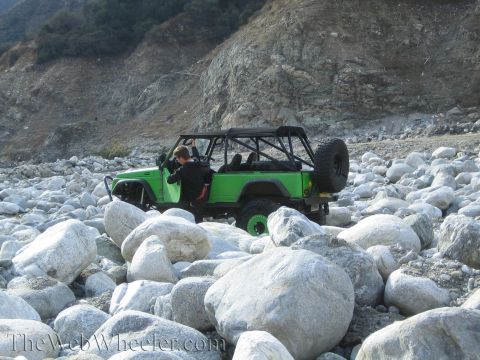 1987 Suzuki Samurai - A person enters a green off-road vehicle on a rocky terrain with a mountain in the background.