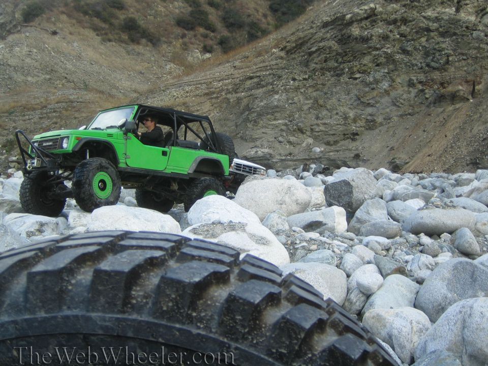 Green off-road vehicle with roll cage driving over rocks in a dry riverbed. Another vehicle is visible behind it