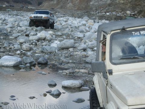 1987 Suzuki Samurai - A beige off-road vehicle, possibly a Suzuki Samurai, drives through a rocky stream with a woman at the wheel.