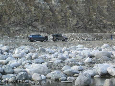1987 Suzuki Samurai - Two SUVs are parked on a gravel road beside a rocky cliff face, with a foreground of large river stones.