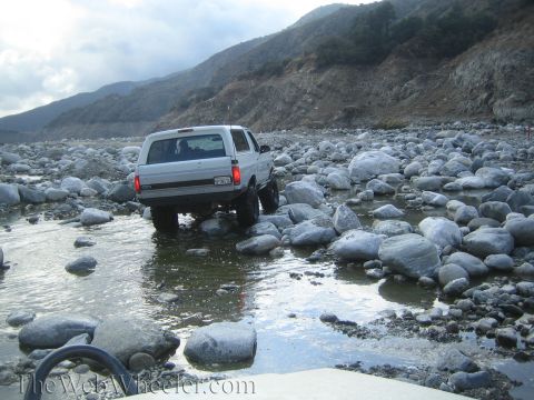 1987 Suzuki Samurai - A white 1987 Suzuki Samurai navigates a rocky riverbed under a cloudy sky.