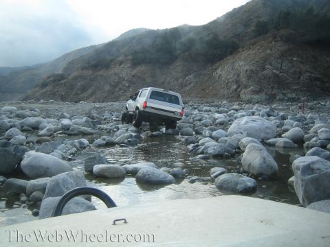 1987 Suzuki Samurai - A white SUV carefully drives through a rocky riverbed, with another vehicle visible in the foreground.